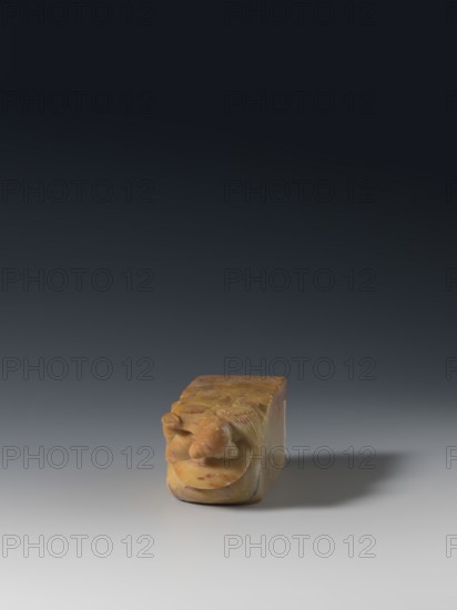 Seal stamp; maker, inscription on the stamp; Chinese; Qing dynasty 1644–1911, 18th–19th century; material: sculpture seal stamp; dimensions: height 9.3 cm, width 3.2 cm, depth 3.1 cm; subject/iconography: Taoism — Immortals, Eight Immortals; function: seal stamp; style: Qing., A small rectangular carved object sits on a smooth surface casting a soft shadow, carved with a rounded decorative element at one end that has curved and layered shapes; the object appears to be made of a marbled material with tones of yellow, ochre, and pale orange with small darker reddish flecks and some lighter cream areas, and the background transitions from dark gray at the top to lighter gray toward the bottom.