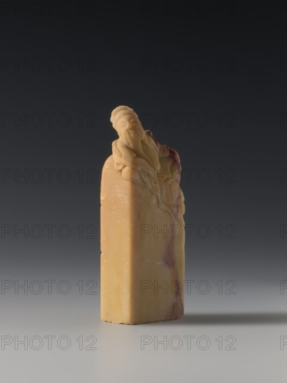Seal stamp, Chinese, Qing dynasty 1644–1911, Qing style, seal stamp depicting sages and officials, 18th–19th century, China; sculpture; height 8.2 cm, width 3.3 cm, depth 1.6 cm., A small carved object stands upright on a smooth surface against a dark gradient background. The object is a vertical rectangular block topped with a sculpted figure that leans forward; the carved details include flowing drapery and rounded facial features. The material shows a warm yellow-beige tone with veins and patches of reddish-brown and subtle lighter areas. The surface displays slight scratches and polished highlights, and the piece casts a soft shadow on the surface beneath it.