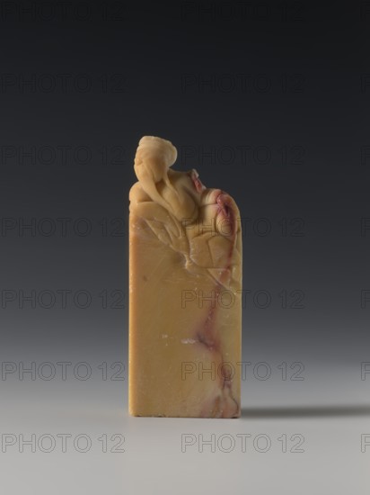 Seal stamp, Chinese, Qing dynasty 1644–1911, Qing style; seal stamp depicting sages and officials; 18th–19th century; sculpture; height 8.2 cm, width 3.3 cm, depth 1.6 cm., A small carved stone object stands upright on a smooth surface against a dark gray background, the stone showing a warm yellow-beige tone with veins and patches of reddish-brown and pale cream; the top is carved into a rounded figure with flowing drapery and a curled feature, the carved area sharing the same yellow-beige color with subtle reddish-brown accents along folds, while the rectangular base is smooth and mostly yellow-beige with faint pale cream marbling and thin reddish-brown streaks.