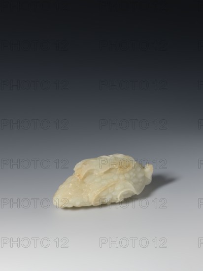 Paperweight - grapevine; maker, pale green jade; sculpture; grapes; writing instrument; Chinese; Qing 1644–1911; 17th–19th century; height 4.0 cm, width 10.3 cm, depth 4.8 cm., A sculpted object made of pale, translucent material with creamy white and light beige tones rests on a smooth surface; the object has an irregular, elongated shape with rounded, overlapping layers and carved, scale-like textures, and it casts a soft shadow on the neutral gray background that gradates from lighter at the bottom to darker toward the top.