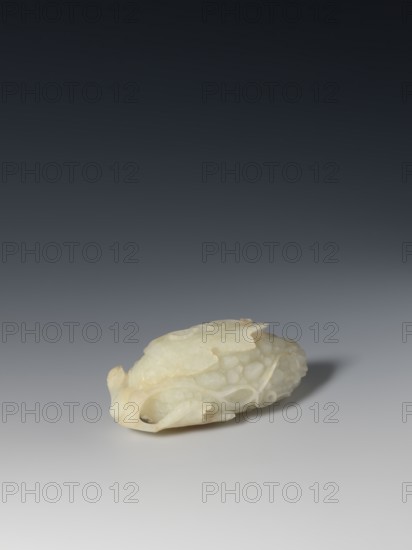 Paperweight - grapevine; maker, Chinese; Qing dynasty 1644–1911; pale green jade; writing instrument; subject: fruit/grapes; sculpture; 17th–19th century; dimensions: height 4.0 cm, width 10.3 cm, depth 4.8 cm., A small carved object rests on a smooth surface, appearing in pale off-white and cream tones with faint warm yellowish areas; the carving has a rounded, elongated shape with intricately carved overlapping scales and flowing, curved motifs, and a few openwork sections that create small negative spaces; the surface shows subtle variations in translucency and texture, with soft shadows beneath it and a darker gray-to-near-black gradient background that shifts from lighter gray near the surface to deep charcoal at the top.