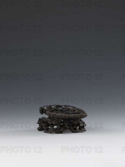 Laozi on base; maker, Qing dynasty 1644–1911, 19th century China; sculpture/figurine of Laozi ca. 550–500 BCE with decoration on base: rocks, pine trees, flowers, lingzhi mushrooms;, Chinese art, Taoism; dimensions — figurine: height 5.2 cm, width 6.3 cm; base: height 7.7 cm, width 6.1 cm., A small dark brown carved wooden stand with an irregular circular top and an openwork base featuring intertwined organic shapes and curved supports, resting on a smooth light gray surface against a gradient background that transitions from light gray near the bottom to dark gray toward the top.