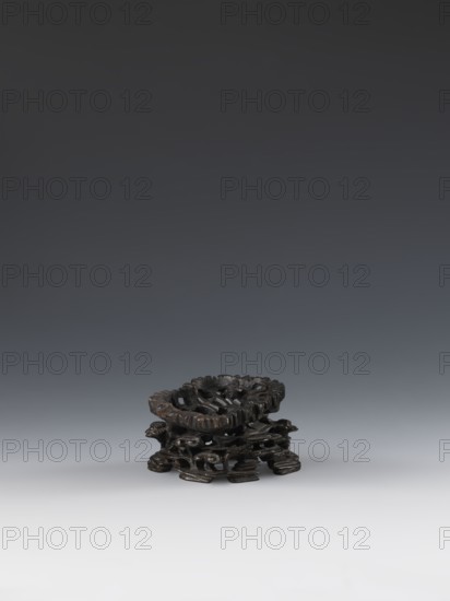 Laozi on base;, deposit provenance; Chinese art; Chinese culture; Qing 1644–1911; Laozi ca. 550–500 BCE; figurine; Taoism; decoration on base: rocks, pine trees, lingzhi mushrooms, flowers; 19th century, China; sculpture; dimensions — figurine: height 5.2 cm, width 6.3 cm; base: height 7.7 cm, width 6.1 cm., A small dark wooden carved object sits centered against a smooth gradient background that transitions from dark gray at the top to light gray at the bottom. The object is a low, round form with an openwork carved top and an intricate base formed by layered organic shapes and carved supports. The wood shows dark brown and almost black tones with some slightly lighter brown highlights on raised areas. The surface appears polished in places and textured in others, with visible carved grooves and rounded contours.