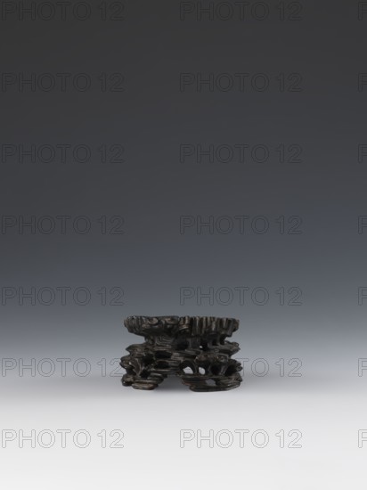 Laozi on base;, deposit provenance; Chinese art; Chinese culture; Qing 1644–1911; Laozi ca. 550–500 BCE; figurine; Taoism; depiction includes rocks, pine trees, lingzhi mushrooms, and flowers — decoration on base: rocks, pine trees, flowers, lingzhi mushrooms; 19th century; China place of origin; sculpture; dimensions: figurine height 5.2 cm, width 6.3 cm; base height 7.7 cm, width 6.1 cm., A small dark brown carved object with an irregular, layered form sits near the bottom center of the image against a smooth gradient background that shifts from dark gray at the top to light gray and off-white toward the bottom; the carved object shows complex openwork, textured surfaces, and varied contours, with highlights of slightly lighter brown on some raised edges.