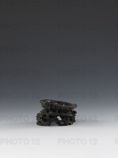 Laozi on base; maker, deposit provenance noted; Chinese art; Chinese culture; Qing dynasty 1644–1911; Laozi ca. 550–500 BCE; figurine; Taoism; decoration on base: rocks, pine trees, lingzhi mushrooms, flowers; 19th century; China place of origin; sculpture; dimensions — figurine: height 5.2 cm, width 6.3 cm; base: height 7.7 cm, width 6.1 cm., A small dark carved object sits near the bottom center of the image against a smooth gradient background that shifts from light gray at the bottom to dark gray at the top; the object appears textured and irregular with layered, organic shapes and rough edges, showing predominantly black and deep brown tones with subtle highlights, and it casts a soft shadow on the lighter surface beneath it.