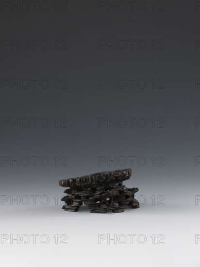 Laozi on base;, Qing dynasty 1644–1911; 19th century; China. Sculpture/figurine depicting Laozi ca. 550–500 BCE with decoration on base: rocks, pine trees, flowers, lingzhi mushrooms. Materials: Chinese art; Taoist subject. Dimensions: figurine height 5.2 cm, width 6.3 cm; base height 7.7 cm, width 6.1 cm., A small carved object sits near the bottom center on a smooth surface, appearing dark brown to black with a glossy finish; it has an irregular flat top with a textured edge and a base made of interlocking organic shapes that form a low, openwork support; the background is a smooth gradient transitioning from pale gray at the bottom to dark gray at the top.
