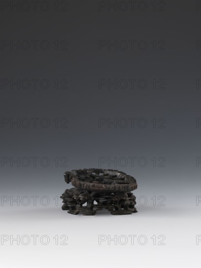 Laozi on base;, Chinese; Qing dynasty 1644–1911; 19th century; taoist subject depicting Laozi ca. 550–500 BCE; sculpture/figurine with decoration on base: rocks, pine trees, flowers, lingzhi mushrooms; materials not specified; dimensions: figurine height 5.2 cm, width 6.3 cm; base height 7.7 cm, width 6.1 cm., A small dark brown carved wooden stand sits near the bottom center of the image, showing a circular top with carved openwork and an intricately carved base with organic, root-like supports; the background above it is a smooth gradient ranging from dark gray at the top to lighter gray toward the bottom, and the surface beneath the stand is a very light gray.