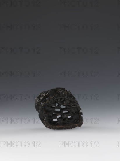 Laozi on base;, 19th century, Qing 1644–1911, China. Figurine and base: sculpture; figurine height 5.2 cm, width 6.3 cm; base height 7.7 cm, width 6.1 cm. Subject: Laozi ca. 550–500 BCE; materials and decoration include rocks, pine trees, lingzhi mushrooms, and flowers. Chinese art; Taoism., A dark carved object sits on a smooth surface against a gradient background. The object is primarily dark brown to black with some lighter brown highlights where the material is worn. It has an openwork, layered structure with irregular holes and carved ridges, and an overall oval or rounded shape. The surface shows textured carving and varied depth, and the base area has a slightly scalloped edge. The background transitions from a deep charcoal at the top to lighter gray toward the bottom, and the surface beneath the object is a pale gray to off-white.