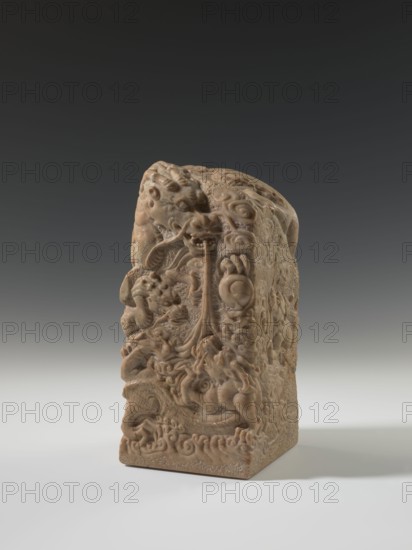 Seal stamp, sculptor/author, carved dragon with a pearl and clouds; Chinese; Qing dynasty 1644–1911, 18th–19th century; sculpture seal stamp; height 26.5 cm, width 12.2 cm, depth 12.5 cm., A carved rectangular stone object with intricate relief work stands on a light surface against a dark gradient background. The object is predominantly beige to light brown with subtle variations in tone across its surface. The carvings depict swirling, rounded forms and a group of stylized animal figures with curved limbs and textured bodies, including coiled shapes and flowing, ribbon-like elements. The top of the object has a rounded, irregular profile with carved details continuing over the edges. The base shows a band of repeating, wave-like patterns and the surface exhibits areas of smoothness and rough, pebbled texture. Shadows fall to one side, creating a soft contrast between the object and the background.