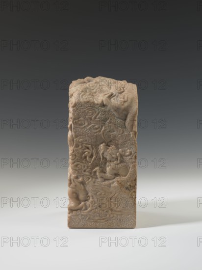 Seal stamp, unknown creator; sculptor, Krieg doctor inscription; Chinese seal stamp depicting a dragon with a pearl amid clouds; iconography: dragons; culture: Chinese; Qing dynasty 1644–1911; dated 18th–19th century; material: sculpture; dimensions: height 26.5 cm, width 12.2 cm, depth 12.5 cm., A vertical rectangular carved stone object with a textured surface and intricate bas-relief depicting swirling cloud-like forms and stylized animal figures, shown standing on a flat surface; the stone has an overall warm beige tone with areas of darker brown and subtle variations, and the background is a smooth gradient from dark gray at the top to light gray near the bottom.