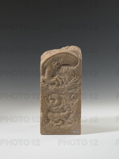Seal stamp, Krieg doctor inscription/ownership; Chinese; dragon with a pearl; dragons iconography; clouds; Qing dynasty 1644–1911; 18th–19th century; sculpture; seal stamp; height 26.5 cm, width 12.2 cm, depth 12.5 cm, A carved rectangular stone object stands upright against a gradient background, showing a predominantly beige and light brown surface with darker brown accents in the carved recesses. The front face displays intricate relief carvings of swirling, organic shapes and coiled forms, with smoother raised areas and textured, stippled background sections. The top edge is rounded and also carved with flowing, curved motifs. The lighting creates soft shadows that emphasize the depth of the carvings, and the base rests on a pale surface that transitions from light gray to white. The visible colors include beige, light brown, darker brown, gray, and white.
