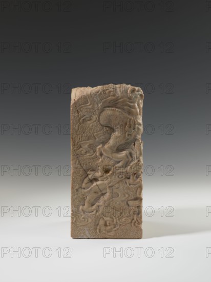 Seal stamp, carved seal showing a dragon with a pearl amid clouds; material: sculpture, Chinese, Qing dynasty 1644–1911, 18th–19th century; dimensions: height 26.5 cm, width 12.2 cm, depth 12.5 cm., A rectangular carved object standing upright on a flat surface, showing a relief design of flowing, sinuous shapes and stylized animal forms on its front face; the material appears to be stone or a stone-like substance with a textured, slightly rough surface and visible cracks and chips; colors visible include shades of beige, tan, light brown, and darker brown accents, set against a neutral background that transitions from light gray at the bottom to dark gray at the top.
