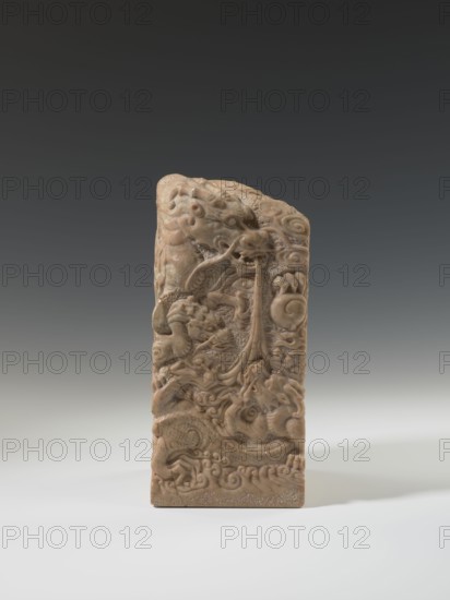 Seal stamp, carved sculpture of a dragon with a pearl and clouds; Chinese, Qing dynasty 1644–1911, 18th–19th century; material: seal stamp sculpture; dimensions: height 26.5 cm, width 12.2 cm, depth 12.5 cm; iconography: dragons, clouds., A carved vertical stone object stands on a pale surface against a dark-to-light gray gradient background, showing a warm beige to light brown color with subtle darker veins. The front face displays an intricate relief of swirling organic forms and sinuous creatures, including curling limbs, rounded eyes, and textured scales, with flowing tendril-like elements and wave-like motifs at the base. The top is irregular and rounded with carved raised patterns, while the sides and base appear smoother and slightly darker. Overall the visible colors are beige, light brown, darker brown accents, and gray tones in the background and surface.