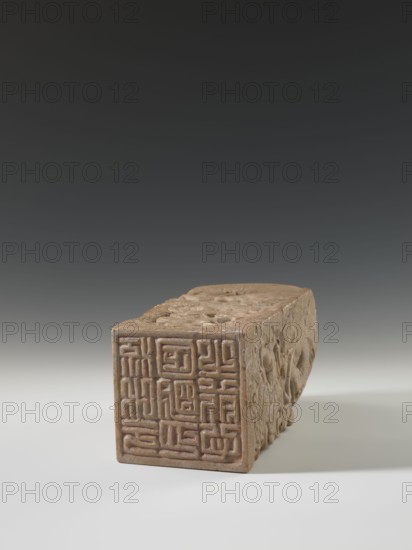 Seal stamp, sculptor; doctor Krieg; Chinese seal stamp depicting a dragon with a pearl amid clouds; Qing dynasty 1644–1911, 18th–19th century; China; sculpture; materials likely stone; height 26.5 cm, width 12.2 cm, depth 12.5 cm., A rectangular carved object rests on a pale surface against a gradient dark-to-light background, appearing in light brown and tan tones with textured carved patterns on its sides and top; the visible end shows a recessed maze-like geometric design, while the long side displays relief carvings with rounded and floral motifs, and the top has shallow carved circular and linear details; shadows beneath and to the right give a subtle three-dimensional effect.