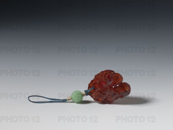 Grape wine pendant, pendant of fossil amber carved with grapes; Chinese, Qing dynasty 1644–1911, 18th century; jewelry; height 3.7 cm, width 3.1 cm., A small carved object made of translucent amber-colored material rests on a smooth surface alongside a short loop of dark blue cord and a round bead of light green stone, with a few small metallic spacer beads near the bead; the background shows a gradient from dark gray at the top to light gray near the bottom.