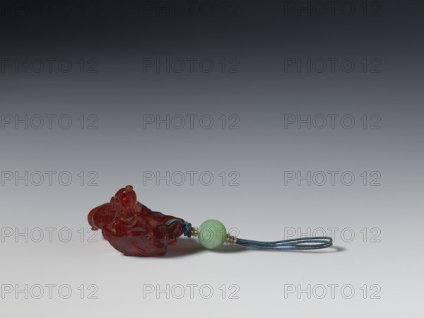 Grape wine pendant, fossil amber; Chinese, Qing dynasty 1644–1911; 18th century; material: amber; subject: grapes; function: jewelry pendant; dimensions: height 3.7 cm, width 3.1 cm., A small carved translucent red object rests on a pale surface, connected by a blue braided cord to a smooth round green bead, with small pale spacer beads between the green bead and the cord; the background transitions from light gray near the surface to dark gray at the top.