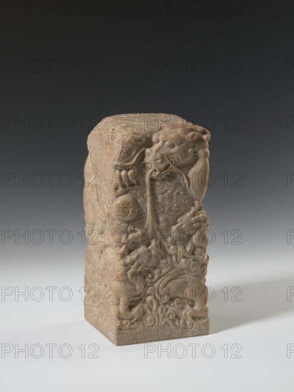 Seal stamp, Chinese, Qing dynasty 1644–1911, 18th–19th century. Sculpture seal stamp depicting a dragon with a pearl; carved in stone, height 26.5 cm, width 12.0 cm. Icons: dragons; style: Qing., A rectangular carved stone object with intricate raised reliefs on its visible faces sits on a pale surface against a dark gradient background; the stone has a warm beige to light brown color with variations and a slightly glossy, smooth appearance in places and rougher texture elsewhere, and the carvings depict swirling organic forms, stylized animal heads with open mouths, curling tendrils, and rounded protrusions, creating a dense, layered surface of overlapping motifs that extend from the top edge down the sides.