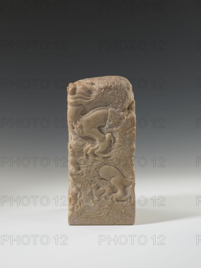 Seal stamp, Chinese art, Chinese culture, Qing 1644–1911, Qing style, seal stamp, dragon with a pearl iconography, dragons, circa 18th–19th century, China, sculpture; height 26.5 cm, width 12.0 cm, A vertical rectangular carved stone object stands on a light surface against a dark gradient background, the stone showing a warm tan to light brown color with areas of lighter flecking and subtle darker veins; the carving depicts flowing, rounded raised forms resembling stylized animals and swirling cloud-like shapes wrapping around the front and sides, with smooth polished areas contrasting with rougher, speckled textures, and the top of the object is rounded and carved with similar organic motifs.
