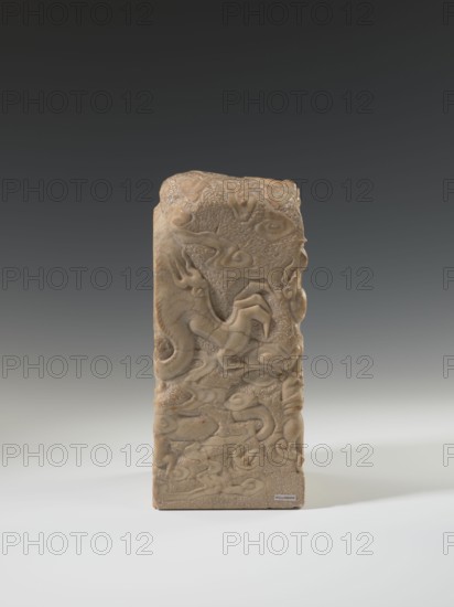 Seal stamp, repossession, Chinese art, Chinese culture, Qing 1644–1911, Qing style, seal stamp with dragon iconography, dragon with a pearl, 18th–19th century, China, sculpture; height 26.5 cm, width 12.0 cm, A carved rectangular stone standing upright with rounded top edges, showing a relief of swirling organic shapes and stylized cloudlike forms across its surface; the stone displays a palette of beige, tan, and light brown with subtle variations and some darker brown veining; the background behind the stone is a smooth gradient from dark gray at the top to lighter gray toward the middle and pale off-white at the bottom; the stone casts a soft shadow on the pale surface beneath it.