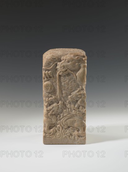 Seal stamp, Chinese art, Chinese culture, Qing dynasty 1644–1911, Qing style, seal stamp, dragon with a pearl iconography, dragons, 18th–19th century, China, sculpture; height 26.5 cm, width 12.0 cm, A tall rectangular carved object stands upright on a smooth surface, showing intricate raised relief work depicting swirling organic forms and animal-like figures; the object has a warm beige to light brown color with subtle variations and slightly darker recesses accentuating the carved details, while the background gradient transitions from dark gray at the top to pale gray near the surface, and soft lighting creates gentle shadows around the base.