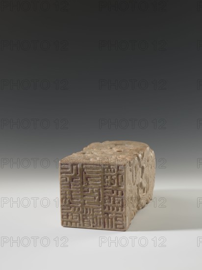 Seal stamp, unknown sculptor; Chinese, Qing dynasty 1644–1911; 18th–19th century. Sculpture of a dragon with a pearl; seal stamp form; carved stone; height 26.5 cm, width 12.0 cm. Iconography: dragons., A rectangular carved object resting on a smooth surface, showing a front face with incised grid-like patterns and a top and side with detailed carved relief; colors visible include beige, tan, light brown, and hints of darker brown and muted reddish tones, set against a gradient background of dark gray to lighter gray and a pale neutral surface.
