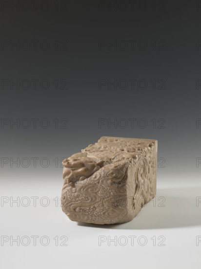 Seal stamp, unknown creator; seal stamp with dragons iconography, dragon with a pearl; Chinese, Qing dynasty 1644–1911, 18th–19th century; sculpture; materials: seal stamp; dimensions: height 26.5 cm, width 12.0 cm; function: seal stamp., A carved rectangular stone block with rounded carved details sits on a smooth surface, showing intricate raised swirling patterns and relief figures on its visible faces; the object appears in shades of beige and light brown with subtle darker brown accents, and the surrounding background transitions from pale gray near the surface to dark gray toward the top.