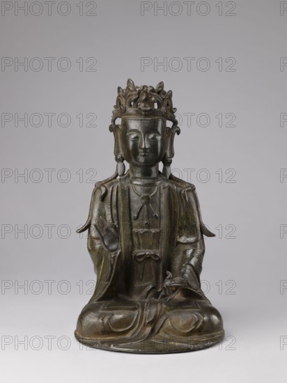 Guanyin;, Chinese; 17th century; China; sculpture, figurine; height 43.0 cm, width 23.0 cm; Chinese Buddhism; bodhisattva; protective deities iconography., A seated bronze statue of a robed figure with a decorative crown, shown against a plain light gray background; the figure has a calm, symmetrical face with closed or lowered eyes, elongated earlobes, and a rounded headpiece with ornate openwork elements. The figure wears layered robes with flowing sleeves and draped folds, and a few hanging adornments at the chest; one hand is raised with fingers forming a gesture while the other rests on the lap holding a small object. The surface shows a dark brown to greenish-brown patina with subtle variations and highlights, and the base on which the figure sits is rounded and slightly worn, matching the overall bronze coloration.