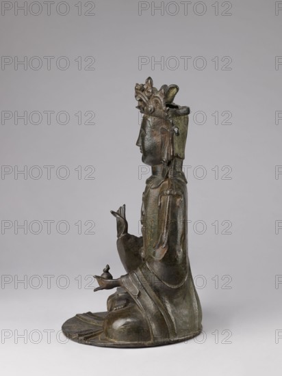 Guanyin, Chinese, 17th century, China, sculpture figurine, bodhisattva and protective deity iconography, Chinese Buddhism; height 43.0 cm, width 23.0 cm., A bronze statue of a seated figure shown in profile against a plain light gray background, with a dark greenish-brown patina and subtle highlights of green; the figure wears a tall ornate headdress with carved decorative elements, has stylized facial features and long earlobes, and wears flowing layered robes with visible folds and rounded drapery; one hand is raised with slender fingers in a gesture while the other hand rests on the lap holding a small rounded object, the figure sits on a rounded base that follows the contours of the robes, and the overall color palette includes dark bronze, green, brown, and muted gray.