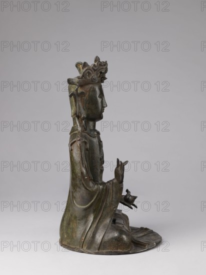 Guanyin, 17th century, China, Chinese art, Chinese Buddhism, protective deities iconography, bodhisattva iconography, sculpture, figurine; height 43.0 cm, width 23.0 cm, A bronze statue shown in profile, seated on a rounded base with draped robes that have smooth flowing folds, with a serene face and closed eyes, wearing an ornate crown with curved decorative elements, an elongated back piece behind the head, and layered jewelry or adornments along the shoulders and chest; one hand is raised with fingers positioned in a gesture while the other rests on the lap holding a small rounded object; colors visible include dark bronze with greenish patina, areas of brown and muted gold highlights, set against a neutral light gray background.