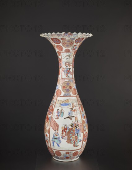 Vase, Arita, Japan; colored porcelain vessel decorated with dragons, figurative scenes, geometric decorations, peonies, chrysanthemums, and other flowers; Meiji period ca. 1870–1880; height 94.0 cm, weight 14.5 kg., A tall, slender porcelain vase with a flared, scalloped rim sits against a dark gray background; the vase has a predominantly white base color covered with decorative panels and roundels in red, blue, gold, and black. The upper flared section features alternating patterned roundels and floral motifs in red, blue, and gold on a white ground, with a scalloped rim edged in gold. The long neck is white with vertically oriented rectangular panels depicting small scenes painted in red, blue, black, and gold, framed by patterned red borders. The bulbous lower body shows a larger rectangular scene with several figures wearing garments in red, blue, and gold, set against architectural elements and a pale landscape; surrounding this scene are circular medallions filled with floral an...