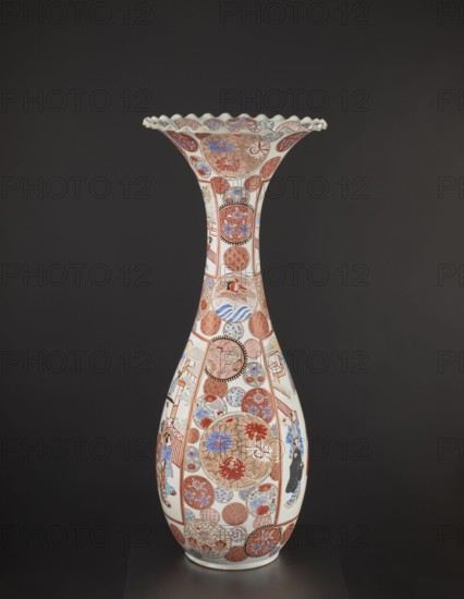 Vase, Japanese; colored porcelain with dragons, figurative scenes, geometric decorations, peonies, chrysanthemums, and other flowers; Meiji period ca. 1870–1880; Arita, Japan; vessel; height 94.0 cm; weight 14.5 kg., A tall, slender porcelain vase with a flared, scalloped rim sits against a dark background; the vase is predominantly white with decorative areas in red, blue, gold, and black forming circular medallions and panels that cover its surface, featuring floral motifs, wave-like patterns, and geometric designs; a few stylized human figures wearing patterned garments appear within some panels; the overall color palette includes white, red, blue, gold, black, and touches of brown, with intricate detailing wrapping the neck and body of the vessel.