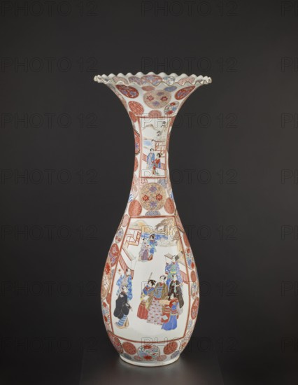 Vase, creator unknown, decorated with dragons, figurative scenes, geometric decorations, peonies, chrysanthemums and other flowers; colored porcelain decorative vessel, Meiji period ca. 1870–1880, Arita, Japan; height 94.0 cm, weight 14.5 kg., A tall, slender porcelain vase with a flared, scalloped rim sits against a dark background; the vase displays a palette of white, red, blue, gold, and black with intricate painted panels and circular medallions; one large panel shows a group of figures wearing patterned robes in hues of blue, red, and gold arranged on a pale background near architectural elements, while another panel higher on the neck depicts a smaller scene with figures and a distant landscape; the body and neck are bordered and interspersed with decorative roundels featuring floral and geometric motifs in red, blue, and gold, and the scalloped rim is edged with gold.
