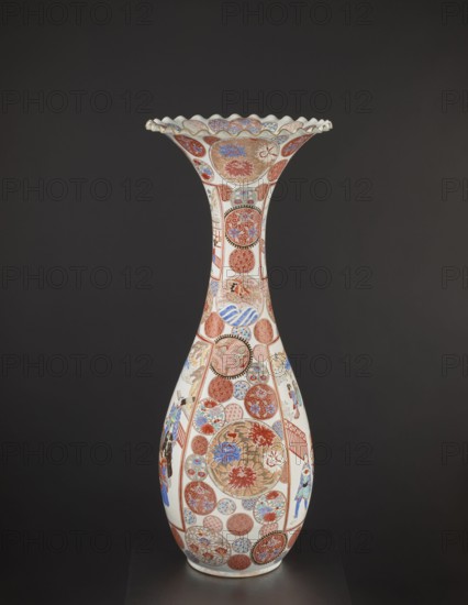 Vase, Japanese, Arita, ca. 1870–1880 Meiji period, 1868–1912. Colored porcelain decorative vessel, height 94.0 cm, weight 14.5 kg. Decoration: dragons, figurative scenes, geometric patterns, peonies, chrysanthemums, other flowers., A tall, slender ceramic vase with a flared, scalloped rim sits against a dark background; the vase has a predominantly white base color decorated with a pattern of many circular and oval medallions in shades of red, blue, gold, and brown, some filled with floral motifs and others with geometric or wave-like designs, and there are a few painted figurative scenes in blue, red, and black on the lower body; the rim edge shows pale blue and gold accents, and the overall palette includes white, red, blue, gold, brown, and black.