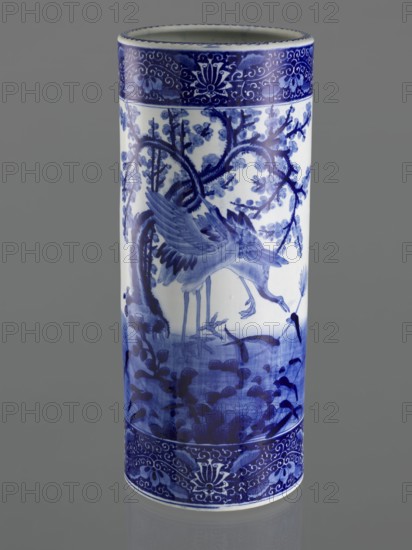 Umbrella vase, ceramics, herons iconography, plum trees, Japanese art, decorative vessel, Meiji 1868–1912, late 19th century, Japan; vessel; height 62.5 cm, diameter 25.5 cm, A cylindrical ceramic vase with a glossy finish shown upright against a plain gray background, decorated in blue and white. The vase displays a detailed painted scene in shades of blue that includes a pair of long-legged birds among branches and foliage, with leafy and floral motifs extending around the body. The top and bottom rims feature ornate border patterns in darker blue with scrolling designs and stylized floral elements. The overall color palette is white with varying blues ranging from deep cobalt to lighter wash tones.
