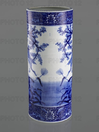 Umbrella vase, ceramics, herons iconography, plum trees, Japanese art, vases, Japanese culture, decorative vessel, Meiji period late 19th century, 1868–1912, Japan; height 62.5 cm, diameter 25.5 cm., A tall cylindrical ceramic vase standing upright with a glossy finish showing a blue and white painted design; the background color of the vase is white with deep blue decorative bands at the top and bottom featuring scrolling floral motifs, and a central scene of blue botanical branches and stylized plants with brushstroke shading; the overall color palette visible is blue, white, and the gray background behind the vase.
