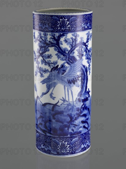 Umbrella vase, ceramics, herons iconography, plum trees, Japanese art, vases, Japanese culture, decorative vessel, Meiji 1868–1912, late 19th century, Japan; vessel height 62.5 cm, diameter 25.5 cm, A tall cylindrical ceramic vase standing upright against a plain gray background, decorated in blue and white. The surface shows a painted scene of birds with long legs and outstretched wings among branches and foliage, rendered in varying shades of blue on a white base. Ornamental bands with floral and scroll motifs in darker blue encircle the top and bottom edges. The overall colors visible are blue, white, and gray.