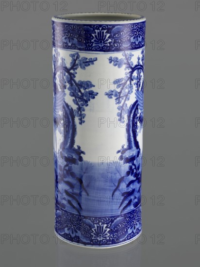 Umbrella vase, ceramics, herons iconography, plum trees, Japanese art, decorative vessel, Meiji period late 19th century, 1868–1912, Japan; height 62.5 cm, diameter 25.5 cm., A tall cylindrical ceramic vase with a glossy white base decorated in a deep cobalt blue. The vase shows a band of intricate floral and scroll patterns near the top and bottom edges, with a central scene featuring stylized branches and foliage rendered in varying blue tones. The blue decoration includes darker outlines and lighter shaded washes, creating a sense of depth on the white surface. The background behind the vase is a neutral gray.