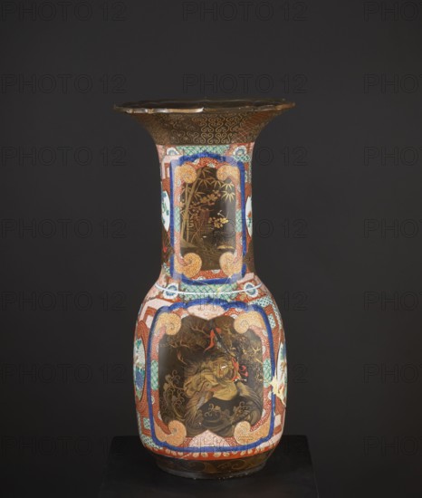 Vase, Japanese, Meiji 1868–1912, c. 1880–1900. High, wide neck with a large flaring cup-like rim; rim with a variegated cut. Decorated with multicolored enamels in the background and in the white reserves; fields covered with black lacquer decorated with gold, red, and gray-blue lacquer; part in flat relief. Motifs: birds, pines, geometric decorations. Colored porcelain vessel, height 79.0 cm, diameter 38.5 cm. Maker and provenance unknown., A tall decorative vase photographed against a dark gray background, predominantly showing colors of black, brown, gold, red, blue, white, green, and orange. The vase has a wide flared rim with a scalloped edge and a dark brown top band decorated with gold swirls. The neck of the vase is narrow and features ornate framed panels with gold-toned scenes on a dark brown background, bordered by patterned bands in blue, red, green, and white with orange scroll motifs. The bulbous lower body of the vase displays a large central panel with a painted figure ...
