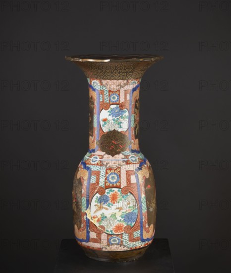 Vase, Japanese art; Japanese culture. Subject: birds and pines. Materials/technique: colored porcelain with multicolored enamels; fields of black lacquer decorated with gold, red, and gray-blue lacquer; part in flat relief. Decoration includes geometric patterns; white reserves. Form/function: decorative vessel—high vase with a wide neck and large flaring cup-like rim; rim with a variegated cut. Style/period: Meiji 1868–1912, c. 1880–1900. Place of origin: Japan. Dimensions: height 79.0 cm; diameter 38.5 cm. Provenance: unknown., A tall, ornate vase is pictured against a dark gray background, showing a flared rim, a narrow neck, and a rounded body. The vase surface displays an intricate patchwork of decorative panels and motifs in a palette that includes red, blue, green, white, gold, brown, orange, and black. Several circular and rectangular panels contain floral scenes with stylized flowers and leaves in blue, orange, green, and white, and a few small birds rendered in muted brown an...