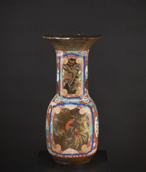 Vase, Japanese, Meiji period 1868–1912, c. 1880–1900. High, wide neck with a large flaring cup-like rim; rim with a variegated cut. Decorated with multicolored enamels in the background and in white reserves; fields covered with black lacquer decorated with gold, red, and gray-blue lacquer; part in flat relief. Subjects include birds and pines; geometric decorations. Colored porcelain, ceramics; decorative vessel vase, height 79.0 cm, diameter 38.5 cm, provenance unknown., A tall decorative vase with a flared rim stands against a dark gray background; the vase features a glossy finish with predominant colors of gold, red, blue, black, green, white, and turquoise. The body shows ornate painted panels framed by red and blue patterned borders, including a large central panel with gold floral motifs and a bird with red and gold plumage, and additional panels with stylized foliage in gold on a black background. The neck and shoulder continue the decorative motif with repeating scroll and ge...