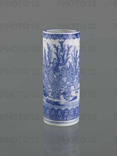 Vessel, Narozno Eckersdorf, Japanese art, Japanese culture, ceramics, figurative scenes, female images, decorative vessel, restitution provenance, 19th century, Japan, vessel, height 39.3 cm, diameter 15.5 cm, A tall cylindrical ceramic vase standing upright against a neutral gray background, predominantly white with intricate blue painted decoration; the blue depicts a detailed scene of figures among tall, slender trees and foliage, with ornate patterned borders near the top and bottom, and a reflected image of the vase visible faintly on the glossy surface below.