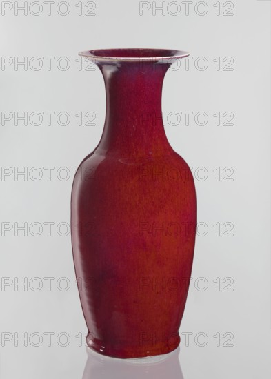 Vase, Chinese, Qing dynasty 1644–1911, 19th century. Large oval ceramic vase with a long wide neck opening, covered with sang de boeuf bull’s blood glaze. Height 58 cm; diameter 24.5 cm., A tall ceramic vase with a flared rim and a narrow neck widening into a rounded body, finished in glossy shades of red and deep crimson with subtle variations and darker red undertones, sitting against a pale gray background and reflected faintly on a glossy surface below.