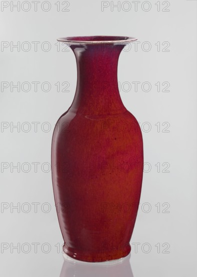 Vase, Chinese, Qing dynasty 1644–1911, 19th century. Large oval ceramic vase with a long wide neck opening at the spout, covered with sang de boeuf bull's blood glaze. Height 58 cm; diameter 24.5 cm., A tall ceramic vase with a glossy red and deep burgundy surface stands against a plain light gray background, showing a flared rim, a narrow neck, a rounded shoulder, and a gently tapering body with subtle darker and lighter red variations and a small reflective highlight near the midsection.