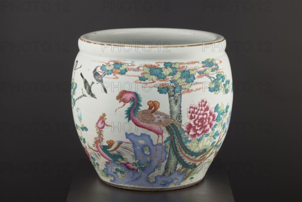 Pool — aquarium., Chinese; Qing dynasty 1644–1911, 19th century. Ceramic colored porcelain vessel decorated with chrysanthemums, lotuses and images of birds; bulging form narrowed at the bottom. Height 41.5 cm; diameter 41.1 cm., A white ceramic jar with a rounded body and a wide open rim, decorated with colorful painted imagery including a pair of stylized birds with long flowing tail feathers in shades of pink, teal, and brown, smaller birds in tones of gray and black perched on branches, and a large peony-like flower in shades of pink and red with green leaves. The painted scene also includes tree trunks and foliage in shades of green, teal, and pale yellow, cloud-like scrolls in pale orange, and rock forms in blue and lavender. The rim shows a thin band of worn brown, and the background behind the jar is a dark gray.