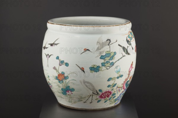 Pool - aquarium, Chinese, Qing dynasty 1644–1911, 19th century. Porcelain colored decoration with chrysanthemums, lotuses and birds. Bulging vessel narrowed at the bottom; height 41.5 cm, diameter 41.1 cm. Function: aquarium/ornamental pool., A rounded ceramic vessel sits against a dark background, predominantly white with painted decorative scenes. The vessel shows a group of birds including cranes with red crowns and a few smaller birds in flight, rendered in shades of black, gray, and white with red accents. There are floral and foliage motifs around the base and sides in colors including teal, blue, green, pink, orange, and purple. A stylized colorful bird or phoenix with bright pink, blue, and orange feathers appears on one side, and several leafy stems and blossoms in green and blue surround the birds. The rim has a weathered brownish edge and the interior appears light grayish white. The piece rests on a dark surface.