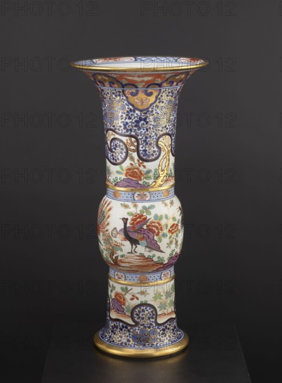 Vase, maker, Japanese; ceramics; colored porcelain Imari; 19th century; floral decorations; phoenixes iconography; lambrequin pattern; decorative vessel/vase; height 66.5 cm; diameter 31.3 cm., A tall ornate vase with a flared rim standing against a dark gray background; the vase displays intricate painted patterns and decorative bands in colors including blue, gold, white, red, green, purple, and black; the top section features dense blue and gold scrolling motifs on a white ground with red and gold accents beneath the flared rim; the central bulbous section shows a painted bird with purple and black plumage standing on a brown rock surrounded by stylized flowers and foliage in red, green, and gold; the lower and upper collars are ringed with patterned bands in blue and white and accented with gold; the base repeats blue and gold scrolling ornament on a white ground with additional floral motifs and a gold trim at the foot.
