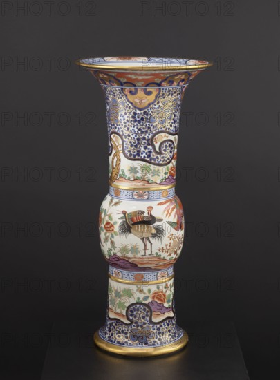 Vase, Japanese, ceramics, colored porcelain Imari, 19th century; floral decorations, phoenixes iconography, lambrequin pattern; decorative vessel/vase; height 66.5 cm, diameter 31.3 cm, A tall decorative vase with a flared rim and a bulbous midsection standing on a round base, decorated all over with intricate painted patterns and motifs in colors including blue, white, gold, red, green, purple, black, and brown; the upper flared section features dense blue scrolling and floral designs with gold accents and red decorative bands near the rim, the central bulbous section shows painted birds amid floral and botanical motifs on a white background with greens and reds, and the lower cylindrical section repeats blue and gold scrolling patterns with floral scenes and a gold band at the base; the vase sits against a dark gray background.