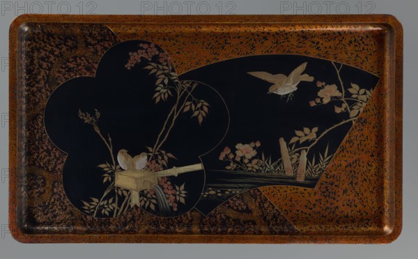 Tray, Meiji 1868–1912, lacquer. With a high vertical rim. Primed wood, black lacquer; decoration dusted with gold, silver, tin, and amaranth paint. Golden-orange background with small flowers, black cartouches with birds among twigs; motifs include kachôga birds and flowers, peonies, water, flowers ornament, fans. 19th/20th century, Japan. Height 3.5 cm; width 50.8 cm; depth 29.4 cm., A rectangular tray with rounded corners shows a speckled rusty orange border surrounding a large black central area shaped with curved insets, featuring painted botanical and bird scenes; within the black area there is a pale beige bamboo water spout with flowing water, two small beige birds perched on the spout, a flying beige bird with spread wings, and several beige and pale pink flowers and leaves on thin brown stems, along with pale green grass and a few beige stumps, all set against the dark background with accents of rusty orange and black speckling on the tray surface.