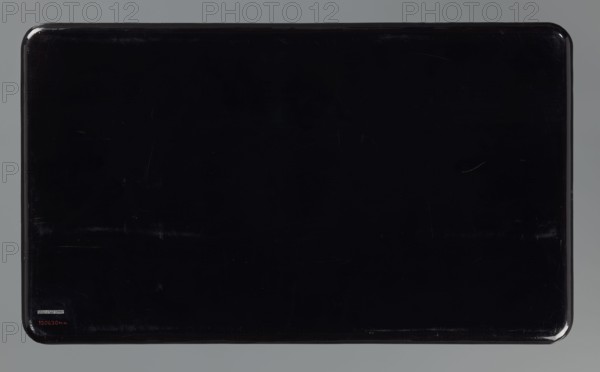 Tray, Meiji period 1868–1912, Japan. Primed wood with black lacquer; decoration dusted with gold, silver, tin, and amaranth paint. High, vertical rim. Golden-orange background with small flowers and black cartouches showing birds among twigs; motif includes peonies, water, and kachôga birds-and-flowers. 19th/20th century. Height 3.5 cm; width 50.8 cm; depth 29.4 cm., classified as a tray., A rectangular, glossy black surface with rounded corners sits against a muted gray background; the black area shows subtle reflections and light scratches, and there is a small square label and a small mark near one corner, while the edges of the rectangle have a slightly darker, reflective rim.