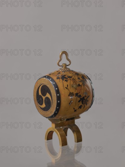 Okimono in the shape of a taiko drum;, possibly Nogawa workshop; ; Japanese, Meiji period ca. 1890–1900; subject: taiko drum with butterflies, insects, plant motifs including paulownia; function: drum-shaped incense burner, vessel; material/technique unspecified; place of origin: Kyoto, Japan; height 15.0 cm, diameter 7.8 cm., An ornate cylindrical object with a rounded top handle sits on a small double-arched stand; the cylinder is primarily golden brown with black floral and leaf decoration painted on its surface, dark circular end panels with a curved triple motif, and darker metal rim accents with small round studs; the stand and handle appear metallic with a brass-like golden color, and the background is a uniform light gray.